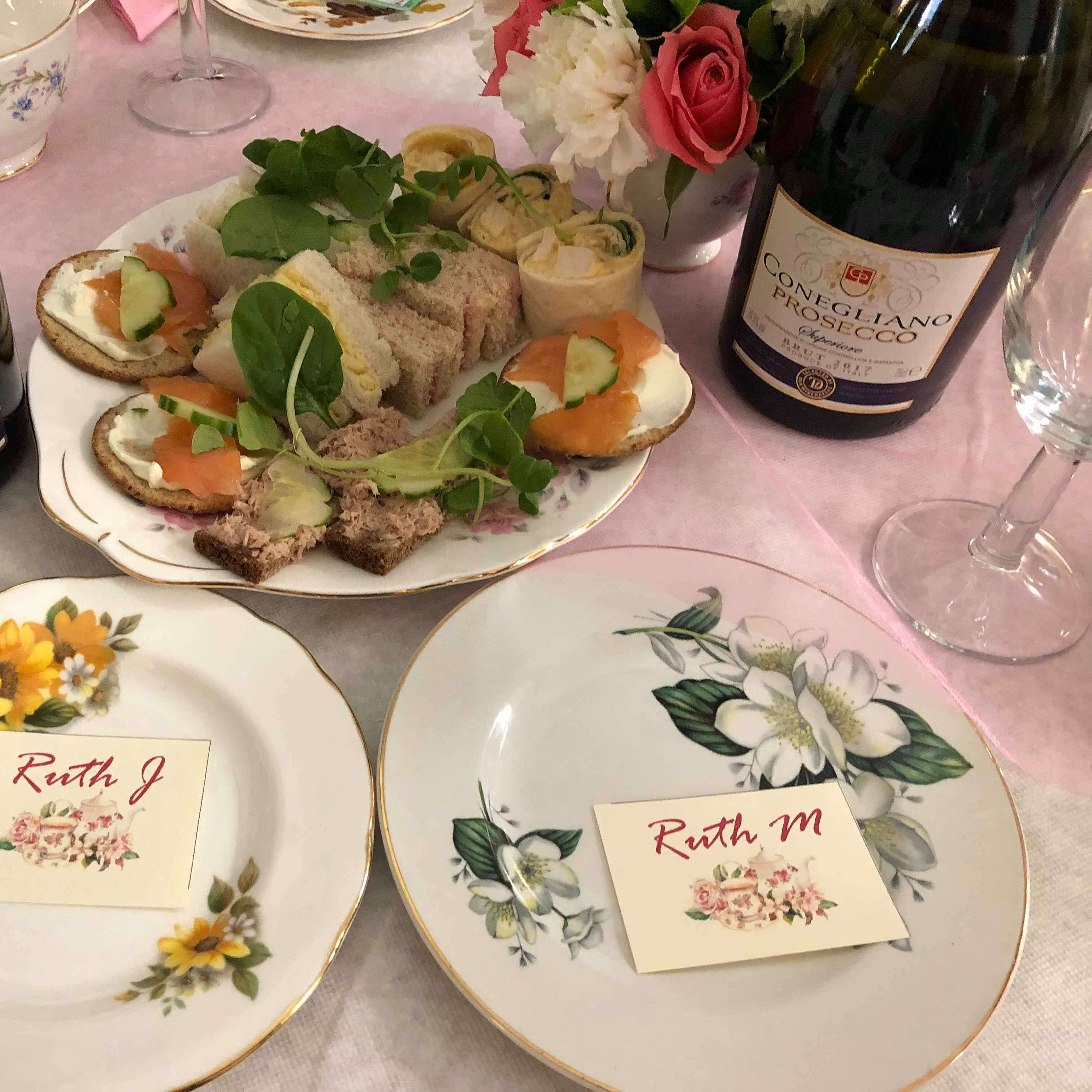 Photo shows a plate of sandwiches, flowers, and Prosecco at an Afternoon Tea Party